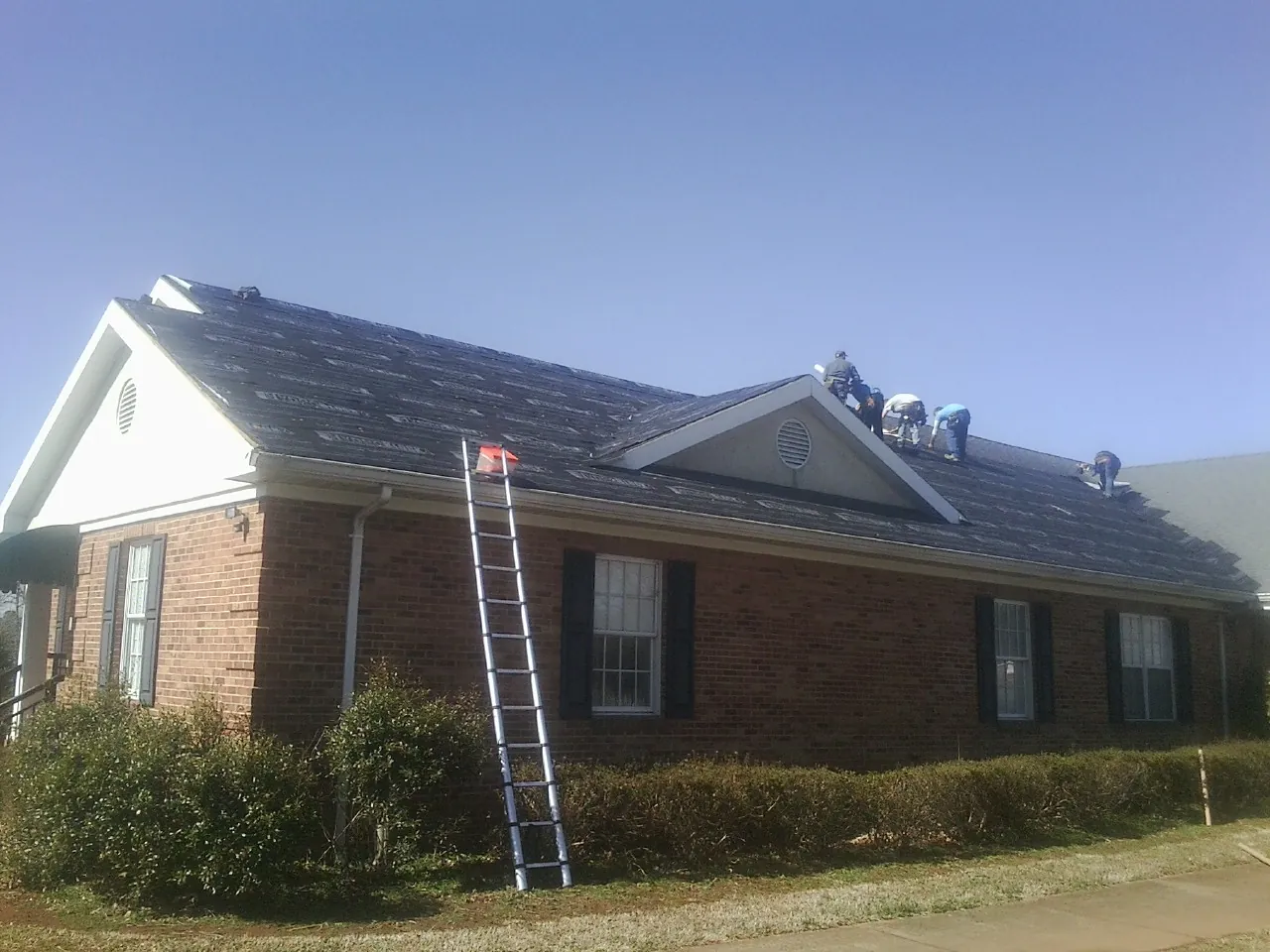 Crew tearing shingles off a brick ranch roof with a ladder set — Emergency Roof Repair work by our crew in Amity
